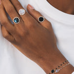 Main d'une femme noire portant trois bagues en plaqué or identiques, mais avec des pierres de couleurs différentes. Chaque bague est dorée et possède une pierre ronde en labradorite naturelle, entourée de zircons naturels sertis. Celle du milieu est en pierre de lune, et la troisième est en onyx noir. Ce sont des bagues élégantes qui attirent l'œil.