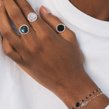 Main d'une femme noire portant trois bagues en plaqué or identiques, mais avec des pierres de couleurs différentes. Chaque bague est dorée et possède une pierre ronde en labradorite naturelle, entourée de zircons naturels sertis. Celle du milieu est en pierre de lune, et la troisième est en onyx noir. Ce sont des bagues élégantes qui attirent l'œil.