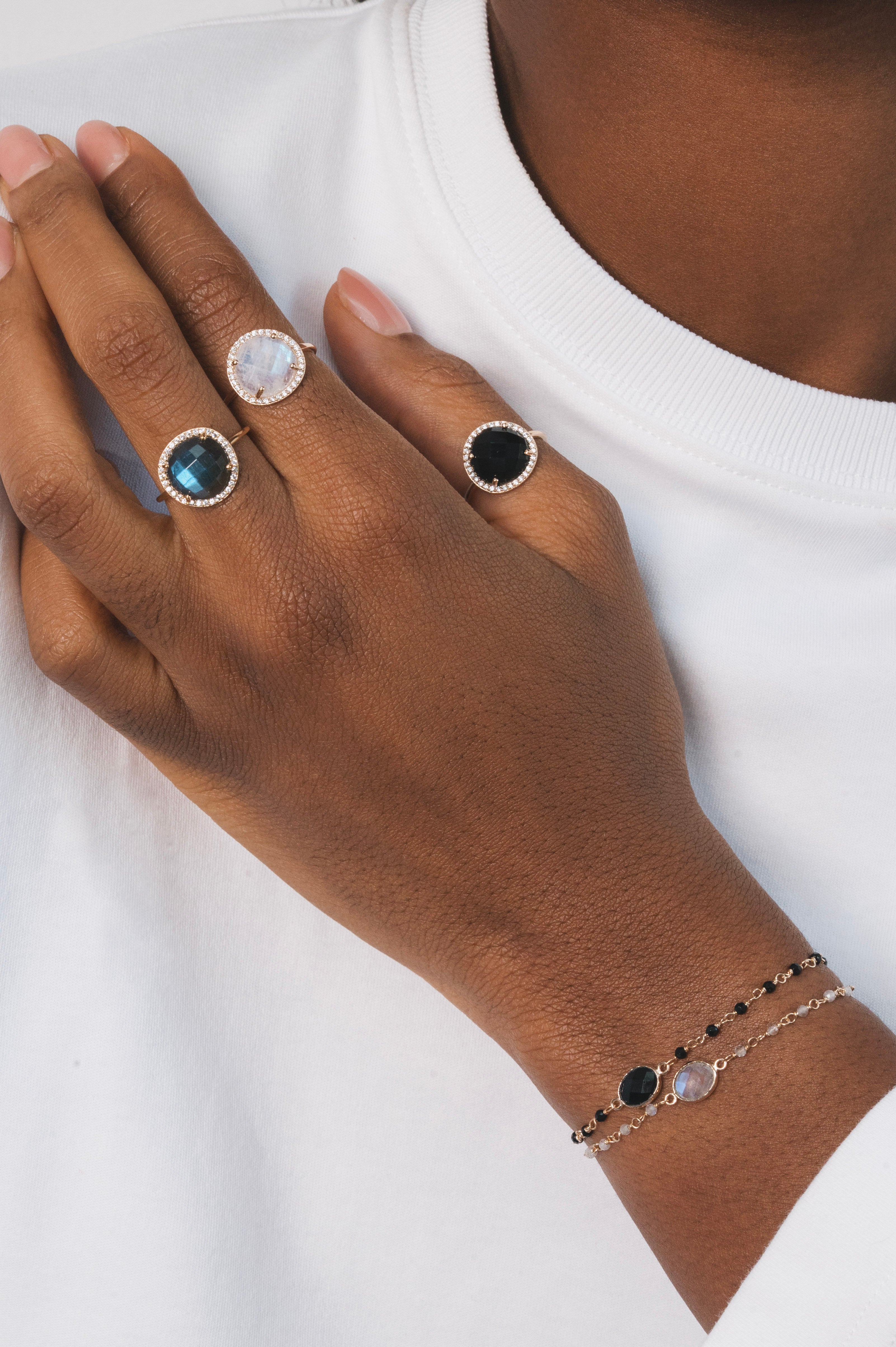 Main d'une femme noire portant trois bagues en plaqué or identiques, mais avec des pierres de couleurs différentes. Chaque bague est dorée et possède une pierre ronde en labradorite naturelle, entourée de zircons naturels sertis. Celle du milieu est en pierre de lune, et la troisième est en onyx noir. Ce sont des bagues élégantes qui attirent l'œil.