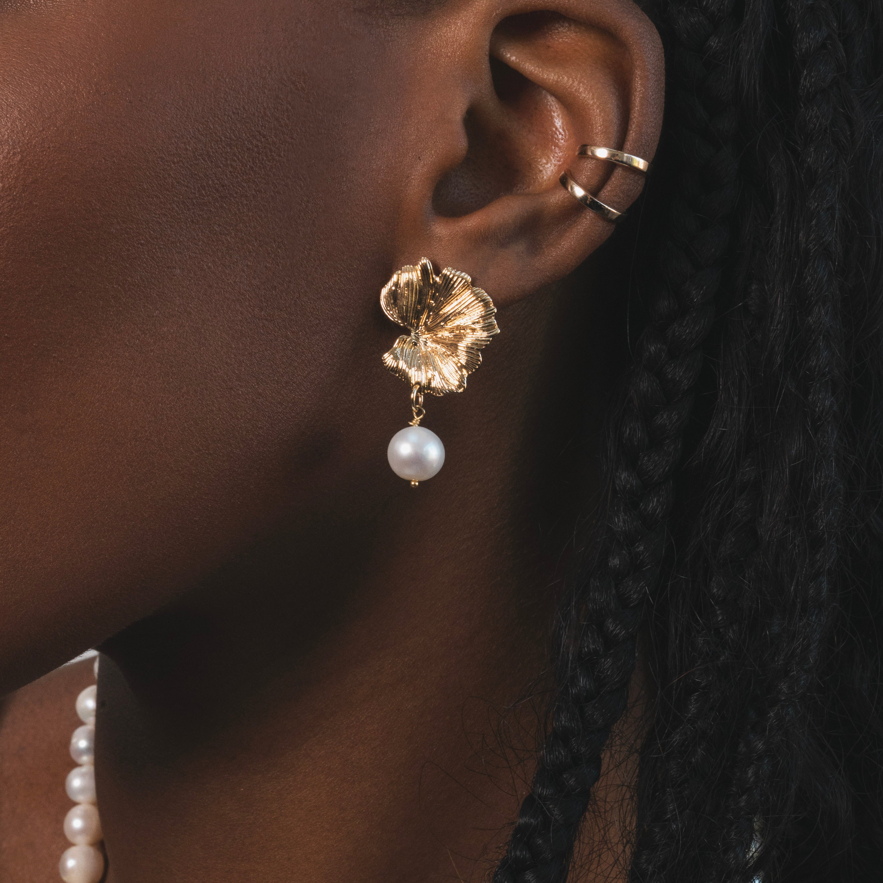 Femme noir qui porte une boucle d'oreille dorée en forme de feuille avec une perle d'eau douces pendantes en dessous. Plus haut sur le cartilage de l'oreille , il y a deux earcuffs dorés. c'est un bijou qui capte la lumière et qui souligne l'élégance naturelle