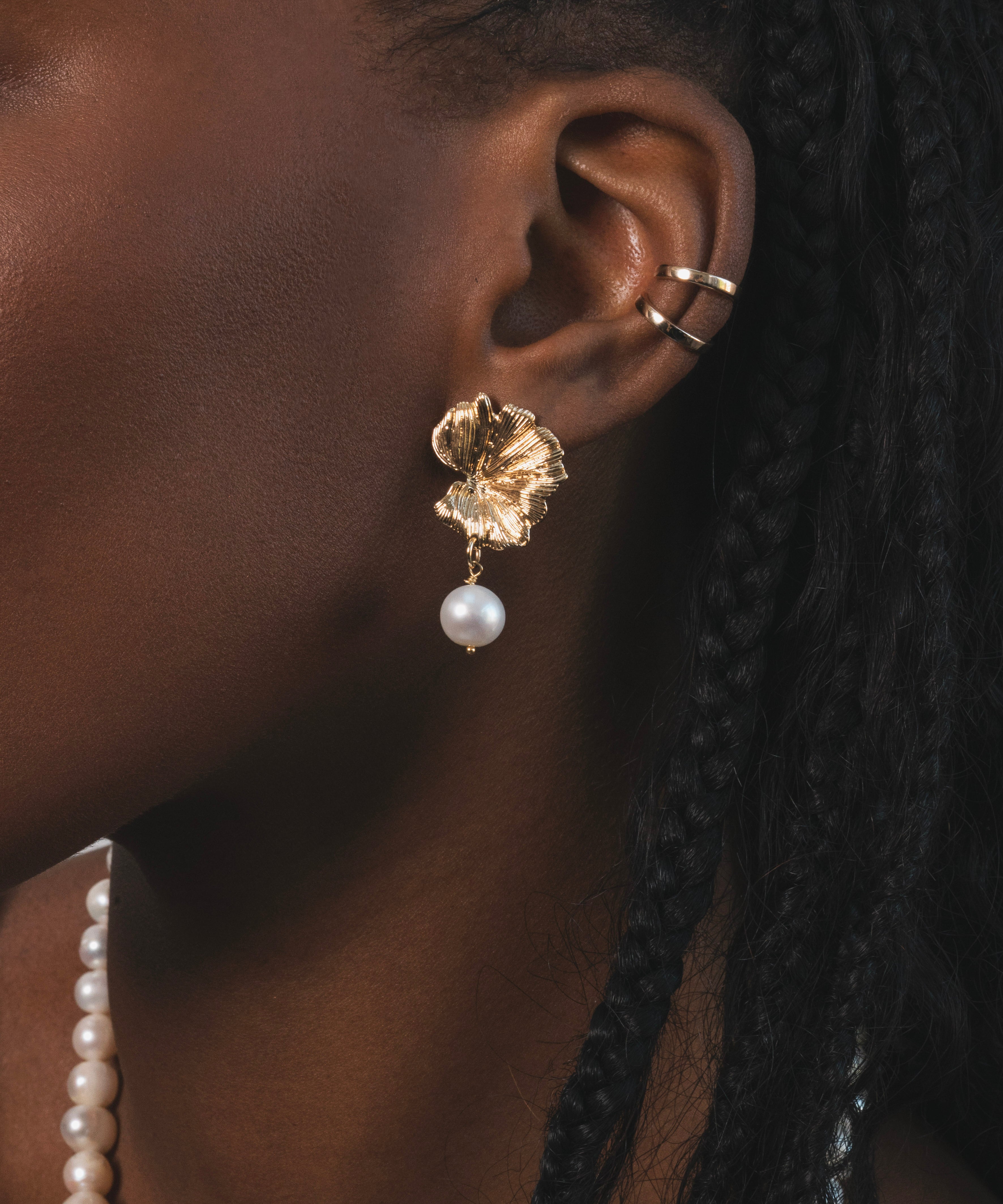 Femme noir qui porte une boucle d'oreille dorée en forme de feuille avec une perle d'eau douces pendantes en dessous. Plus haut sur le cartilage de l'oreille , il y a deux earcuffs dorés. c'est un bijou qui capte la lumière et qui souligne l'élégance naturelle