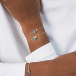 Poignée d'une femme noire, avec deux bracelets en plaqué or en forme de scarabée. Les deux bracelets sont identiques : l'un est en labradorite, une pierre naturelle, et l'autre en amazonite. Ce sont des bijoux lumineux, très légers et raffinés, qui se superposent joliment et apportent une touche originale et colorée ensemble. Ce scarabée représente la protection et le renouveau ; il incarne la résilience, la force et la capacité de se réinventer.