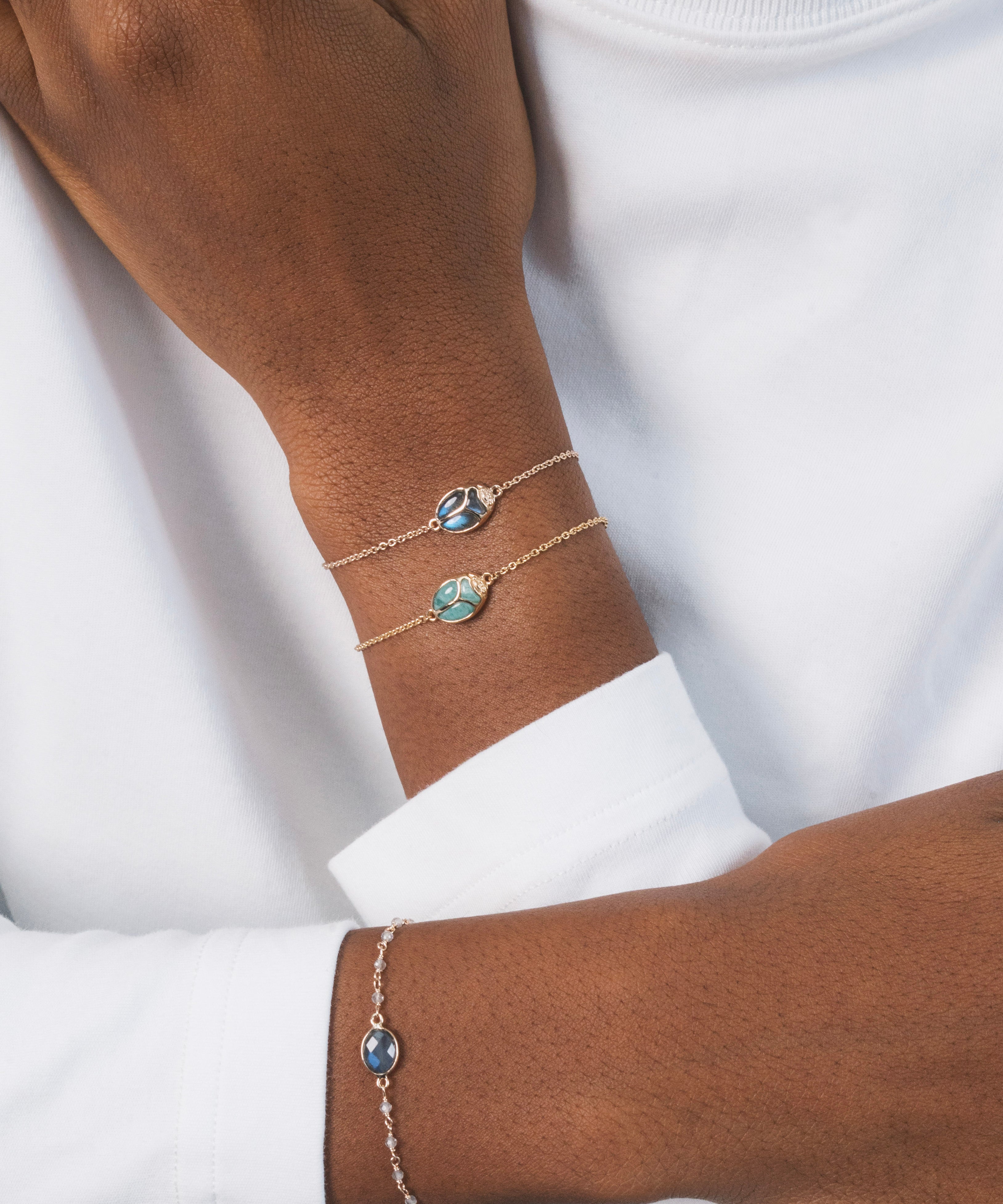 Poignée d'une femme noire, avec deux bracelets en plaqué or en forme de scarabée. Les deux bracelets sont identiques : l'un est en labradorite, une pierre naturelle, et l'autre en amazonite. Ce sont des bijoux lumineux, très légers et raffinés, qui se superposent joliment et apportent une touche originale et colorée ensemble. Ce scarabée représente la protection et le renouveau ; il incarne la résilience, la force et la capacité de se réinventer.