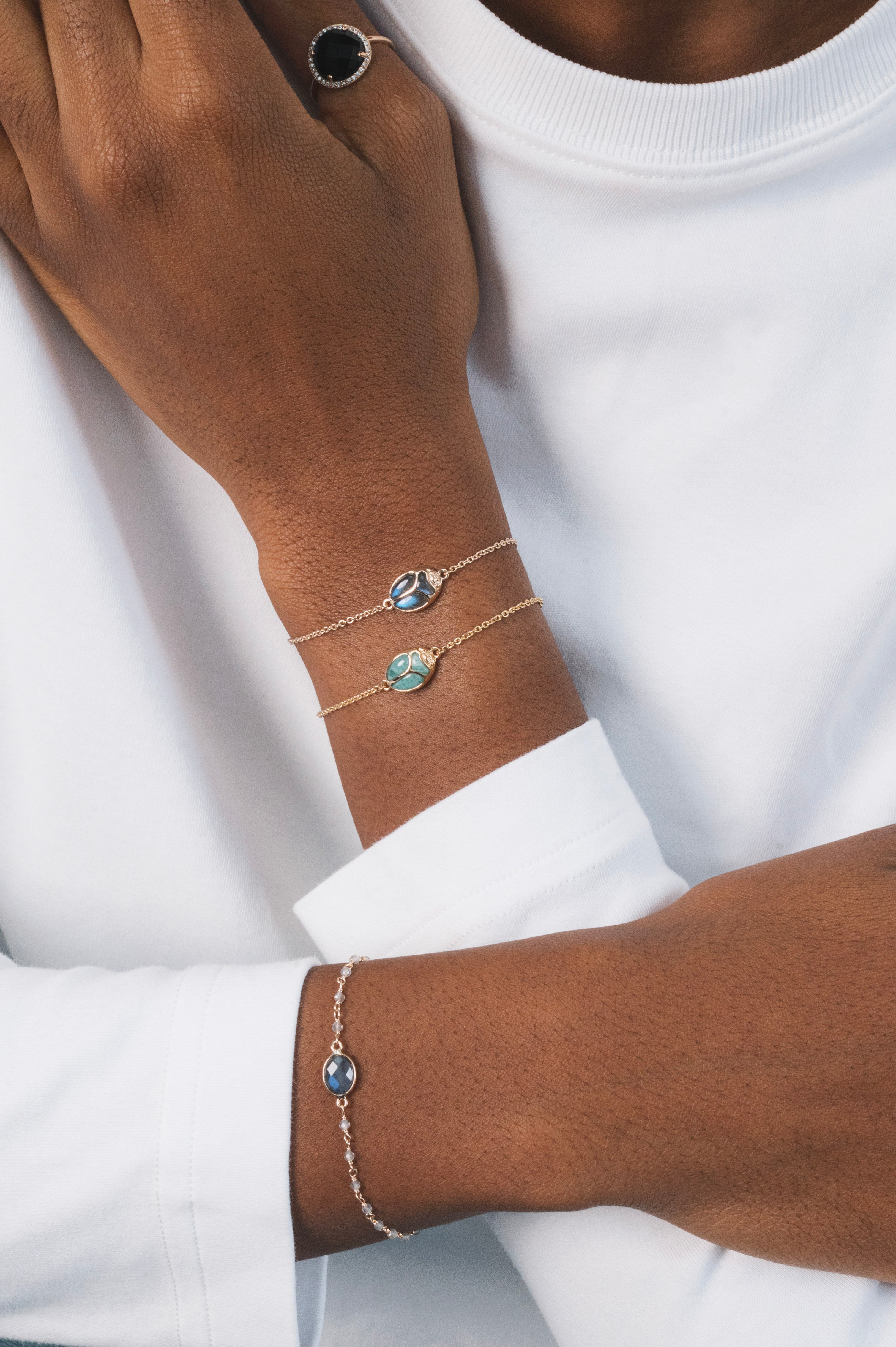 Poignée d'une femme noire, avec deux bracelets en plaqué or en forme de scarabée. Les deux bracelets sont identiques : l'un est en labradorite, une pierre naturelle, et l'autre en amazonite. Ce sont des bijoux lumineux, très légers et raffinés, qui se superposent joliment et apportent une touche originale et colorée ensemble. Ce scarabée représente la protection et le renouveau ; il incarne la résilience, la force et la capacité de se réinventer.