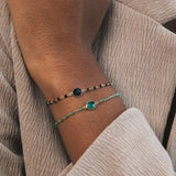 On peut voir un avant-bras et une main portant deux bracelets fins et élégants. Celui du haut est noir et doré, avec une base en plaqué or ornée de petites pierres rondes en onyx noir. Le bracelet du bas est également en plaqué or, avec des pierres en onyx vert.