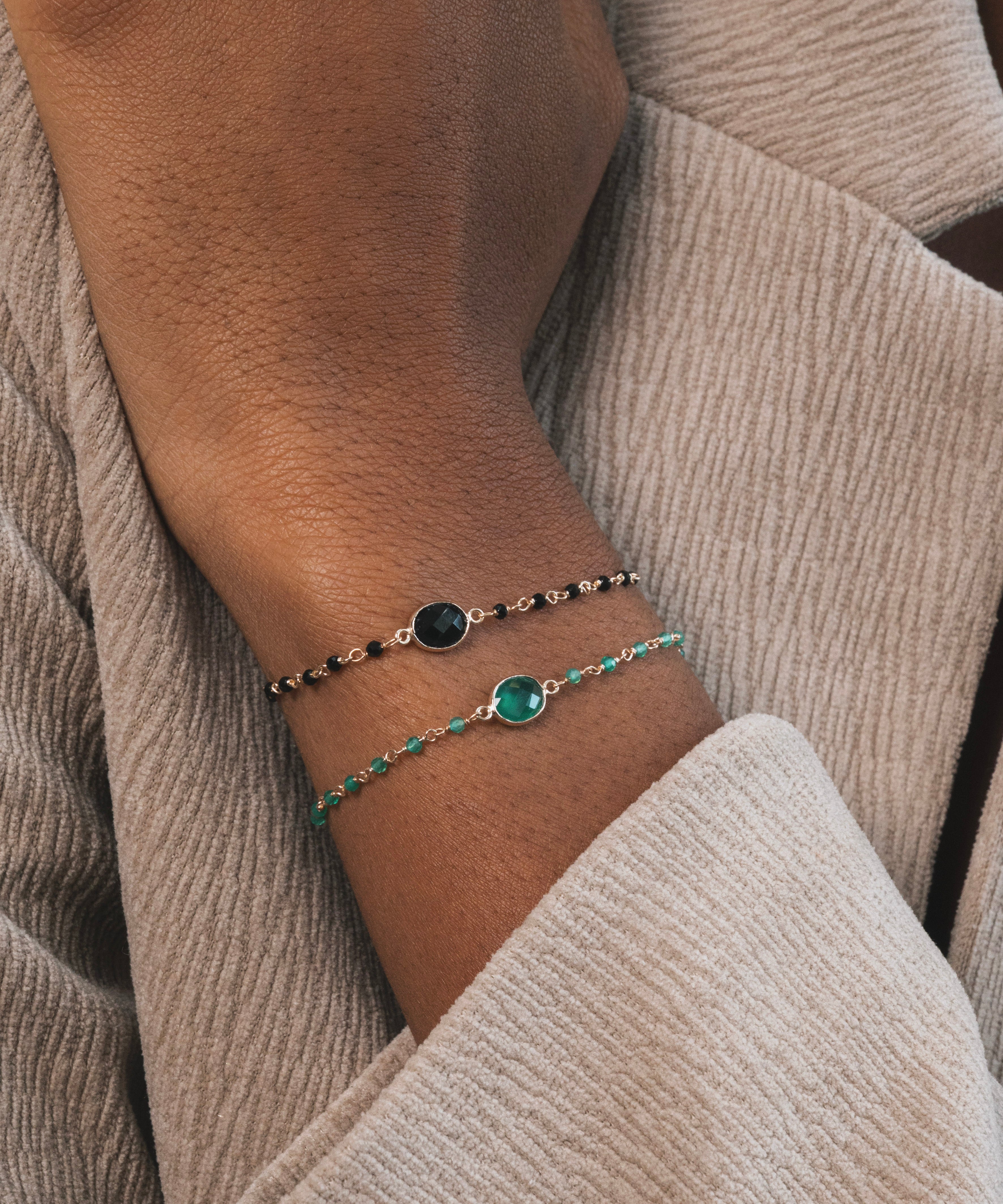 On peut voir un avant-bras et une main portant deux bracelets fins et élégants. Celui du haut est noir et doré, avec une base en plaqué or ornée de petites pierres rondes en onyx noir. Le bracelet du bas est également en plaqué or, avec des pierres en onyx vert.