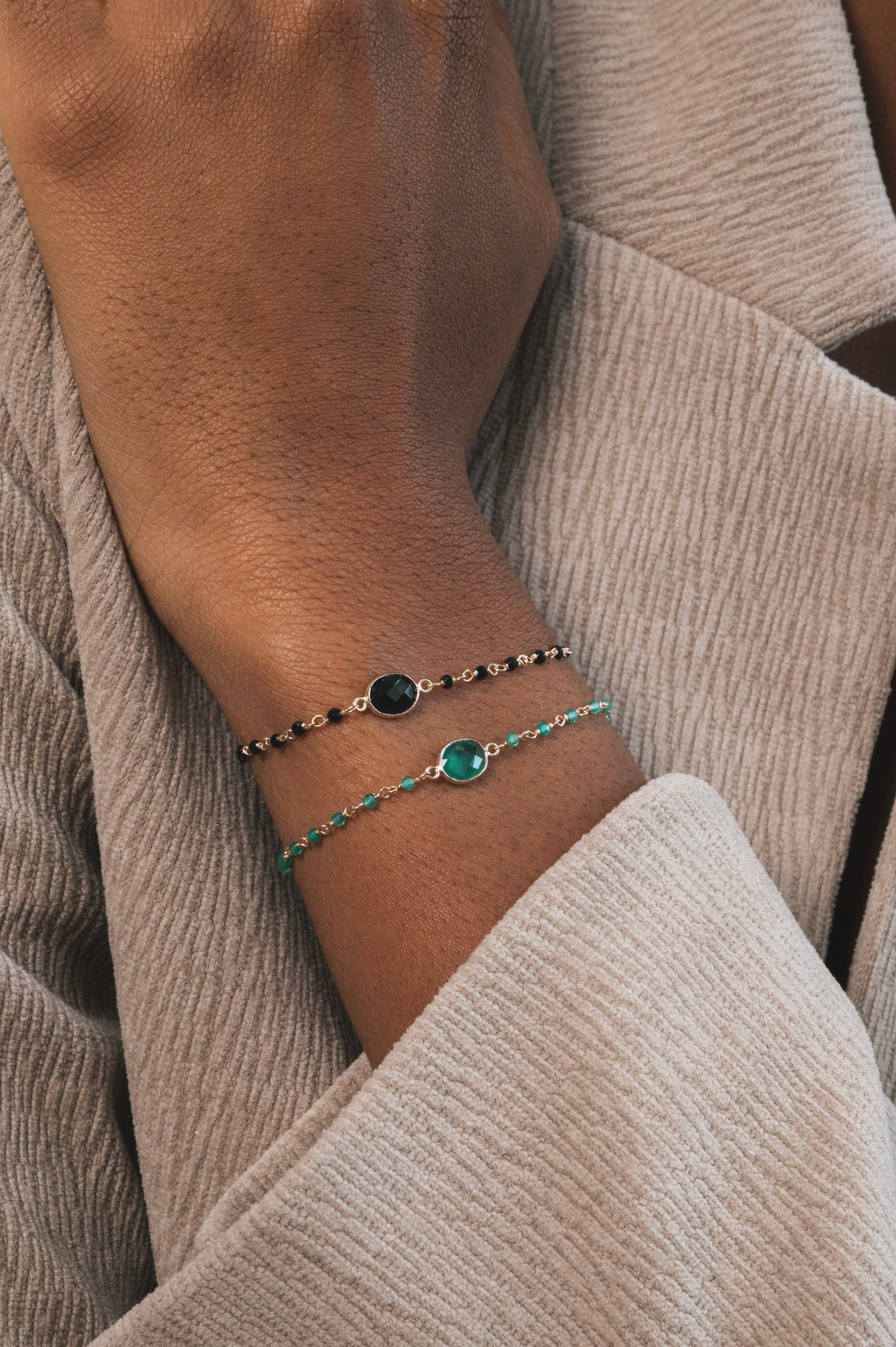 On peut voir un avant-bras et une main portant deux bracelets fins et élégants. Celui du haut est noir et doré, avec une base en plaqué or ornée de petites pierres rondes en onyx noir. Le bracelet du bas est également en plaqué or, avec des pierres en onyx vert.