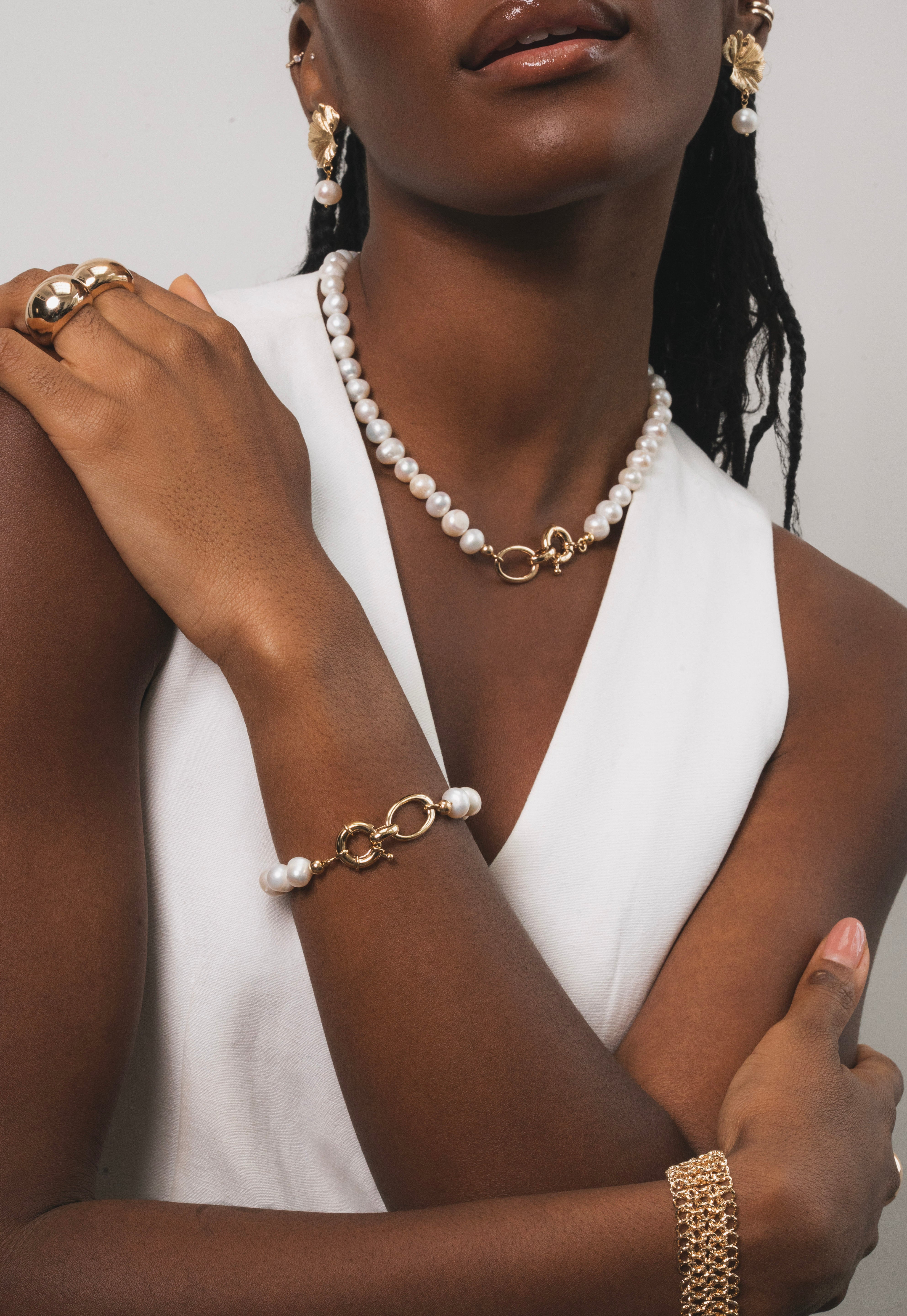 Femme noir qui porte une parrure raffinée qui associe la modernité du doré à la pureté des perles d'eau douces. Chaque piece est pensée pour apporter éclat et élégance, tout en restant facile à porter au quotidien. Le bracelet est raffiné combinant maillons dorés et perles, pour une harmonie parfaite avec le collier. 