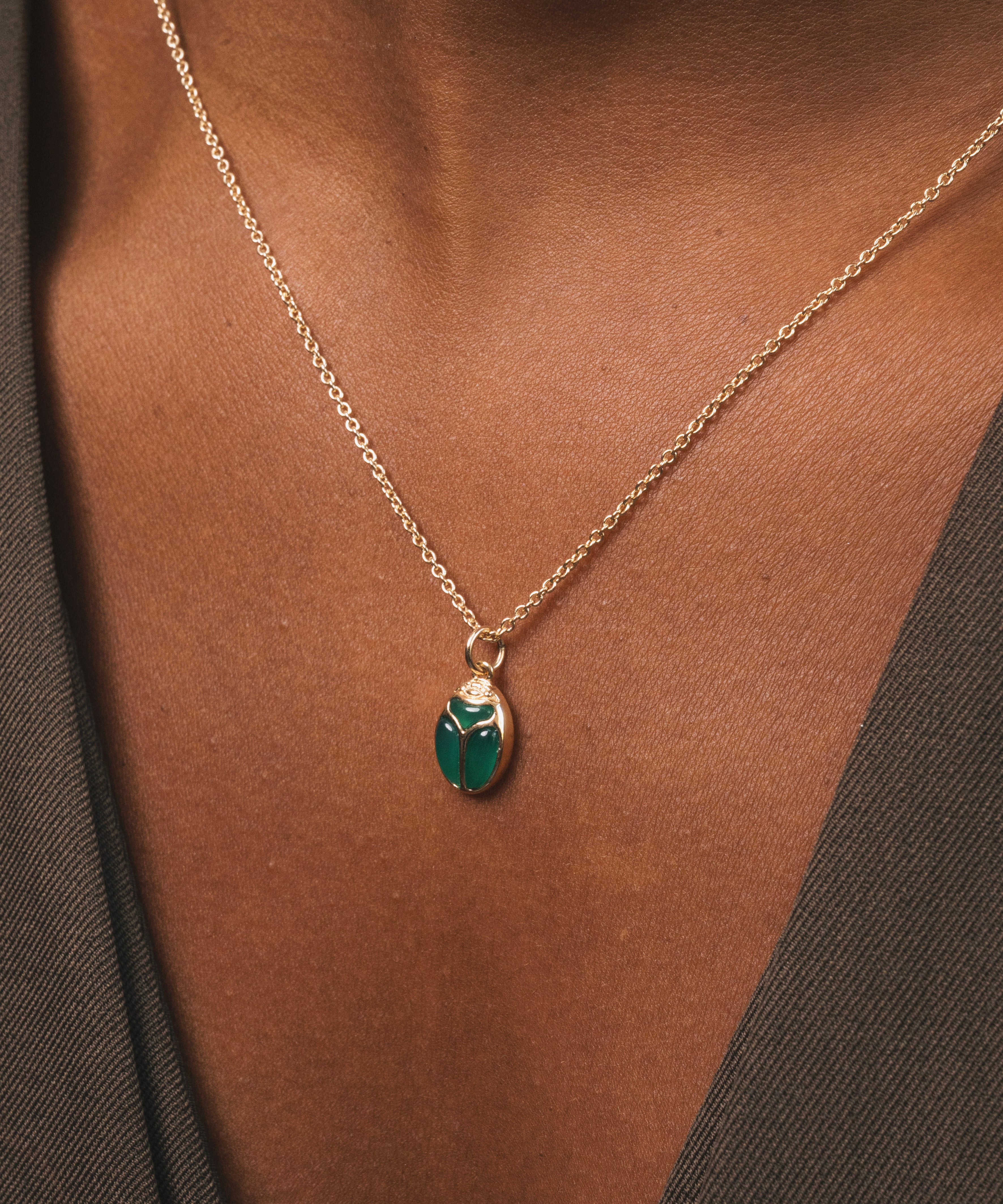 Buste femme noir, qui porte un collier dorée en plaqué or avec comme pendentif  un scarabée en Onyx Vert, une pierre naturelle. Le scarabée a pour symbole de protection, de renouveau, de chance et de prosperité.