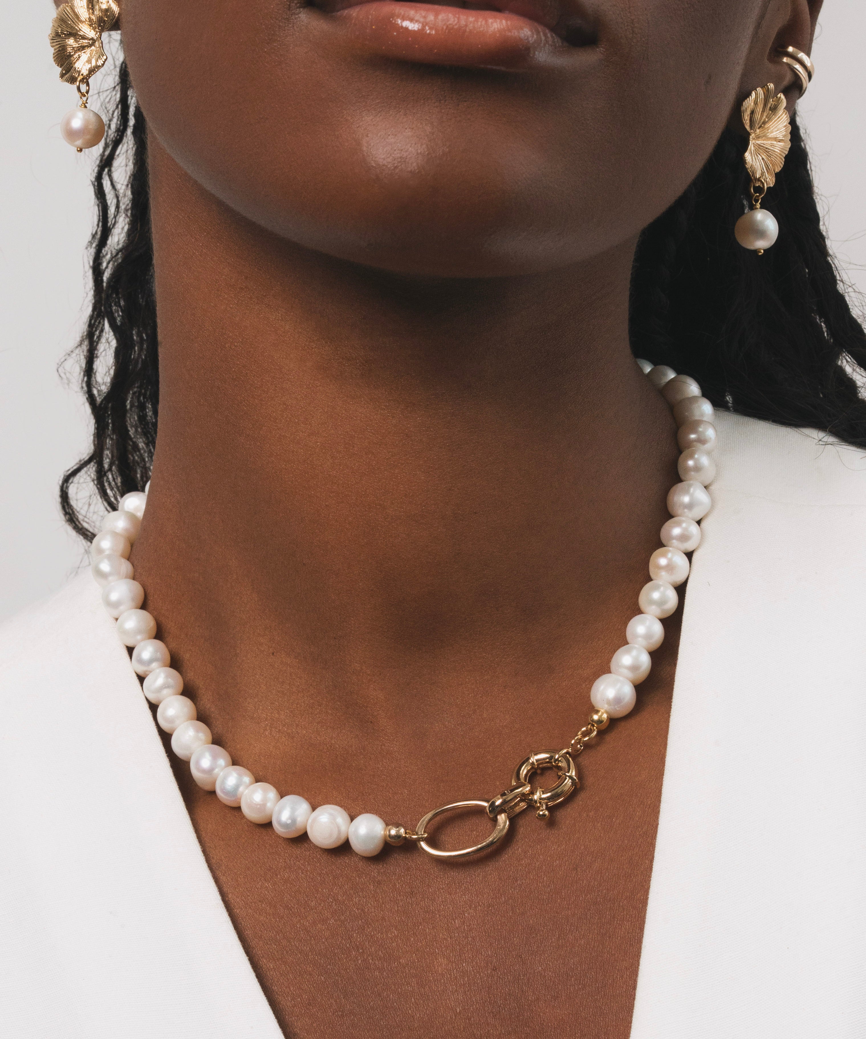 Femme noire qui porte un collier en perles d'eau douce avec des détails dorés en plaqué or. Ce collier a comme particularité un gros fermoir, ce qui fait toute la classe de ce bijou. On peut aussi apercevoir les boucles d'oreilles avec une base dorée en plaqué or ainsi qu'une perle d'eau douce pendante en finition.
