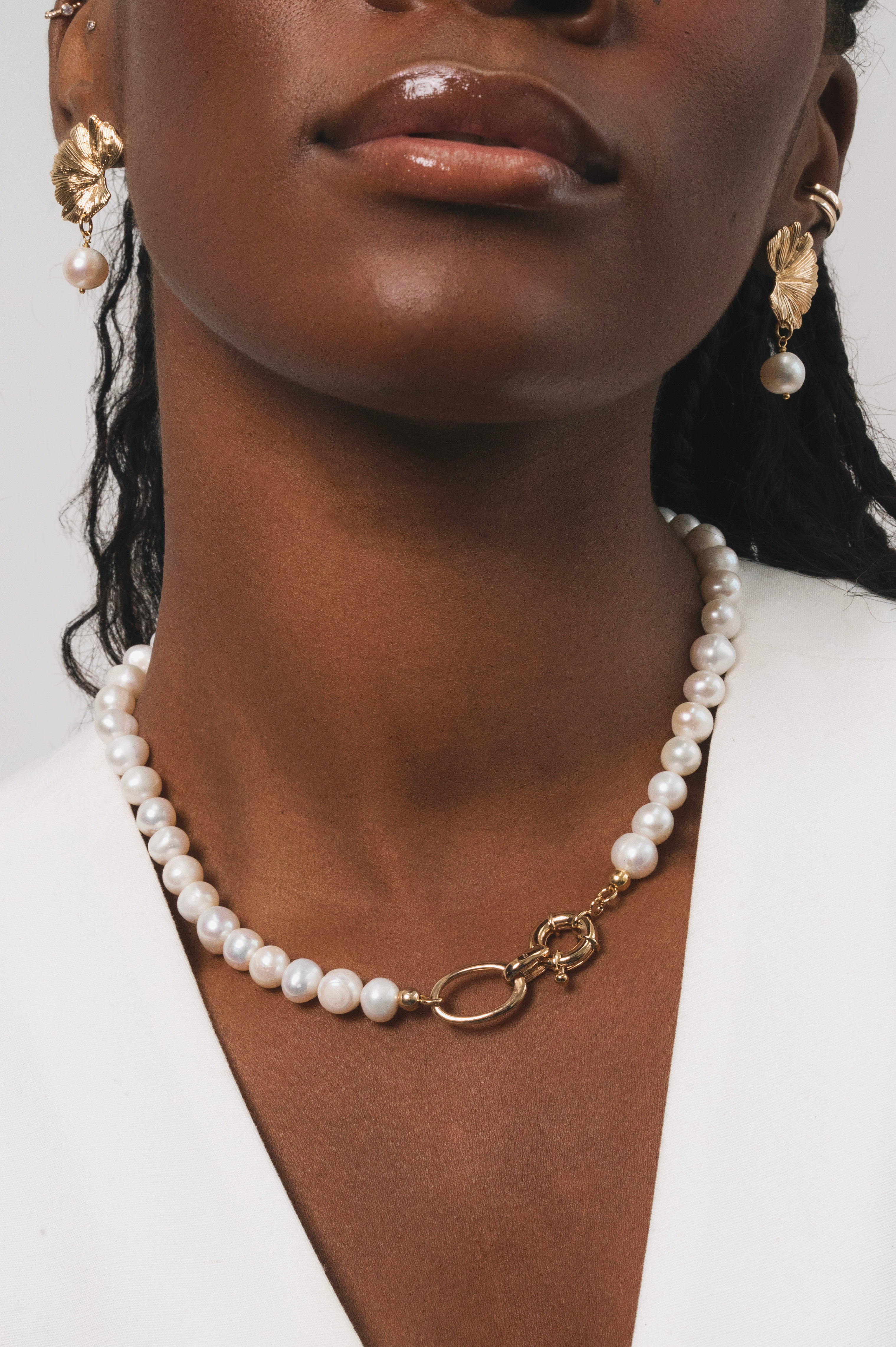 Femme noire qui porte un collier en perles d'eau douce avec des détails dorés en plaqué or. Ce collier a comme particularité un gros fermoir, ce qui fait toute la classe de ce bijou. On peut aussi apercevoir les boucles d'oreilles avec une base dorée en plaqué or ainsi qu'une perle d'eau douce pendante en finition.