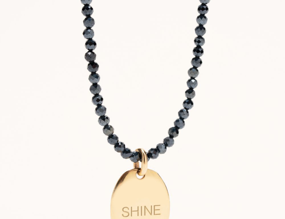 Orphée bijoux collier sautoir de 80 cm porté en double tour de cou avec des perles facettes en hématite bleu gris pétrole et terminé par une médaille ovale bombée sur laquelle il est gravé Shine